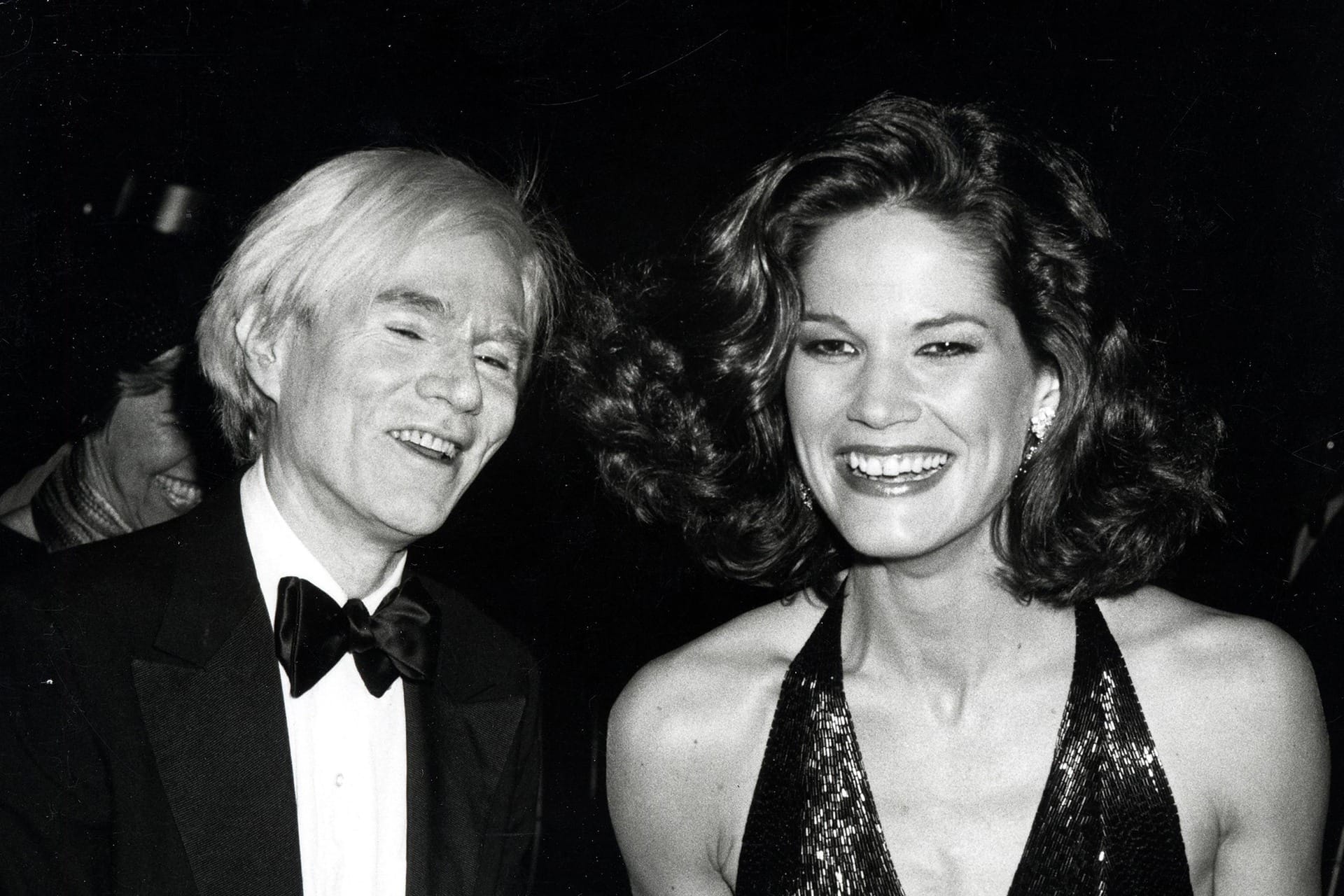A man in a tuxedo and a woman in a sequin dress smiling at a glamorous event.