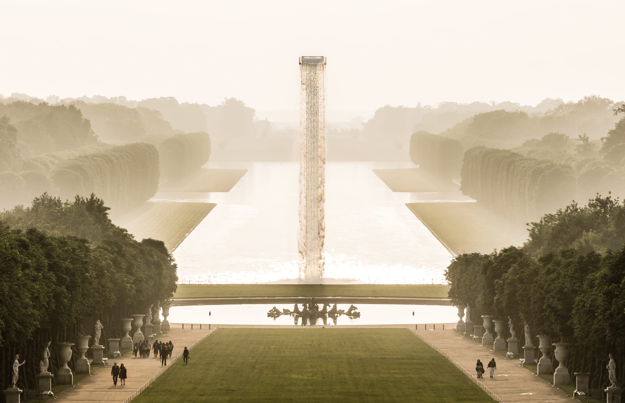 People walking in a garden with a long pathway, tall hedges, and a vertical structure at the end, surrounded by mist.