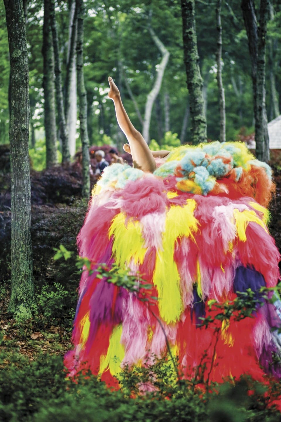 Colorful, furry costume with raised arm in wooded area, vibrant hues of pink, yellow, orange, and blue among green trees.