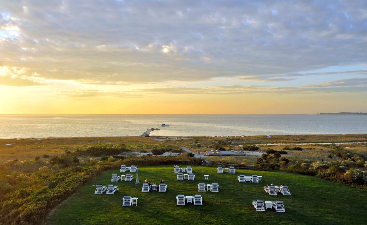 Sunset over a grassy field with white chairs facing a calm ocean and a small pier in the distance.