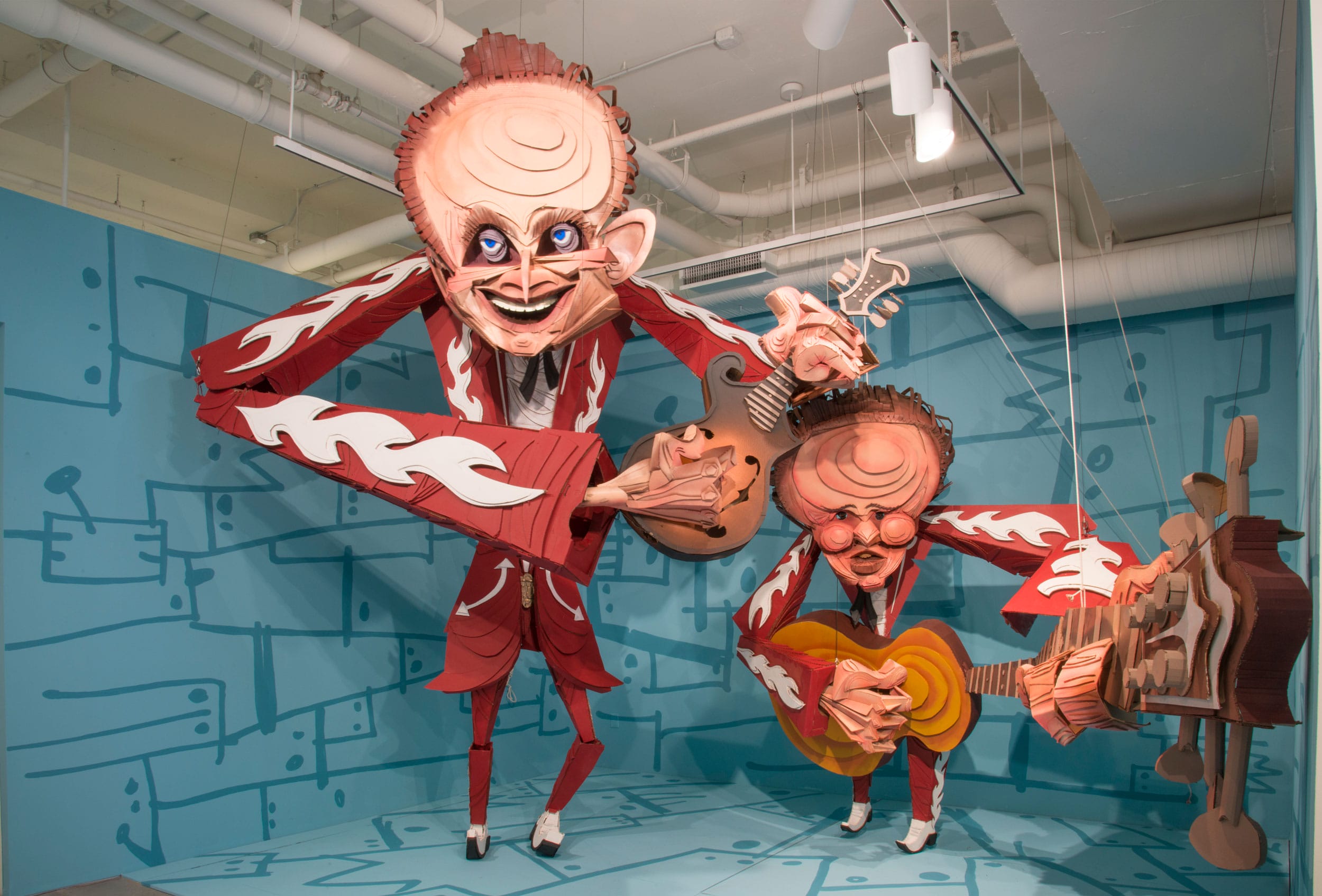 Two whimsical oversized puppets with musical instruments in a colorful room with a blue patterned background.