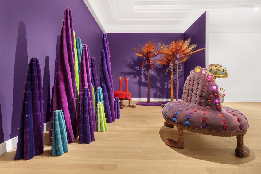 Colorful, whimsical art installation with spiky figures, purple walls, and palm-like sculptures in a gallery setting.