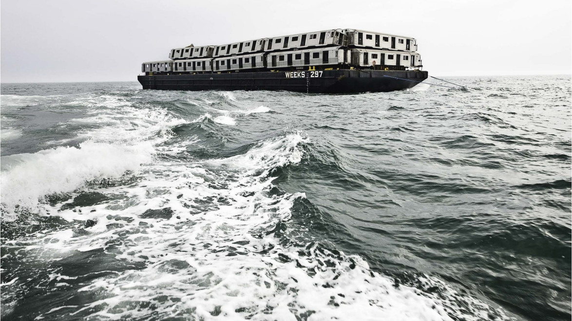 A ship carrying decommissioned subway cars on a vast ocean with a view of the waves and cloudy sky in the background.