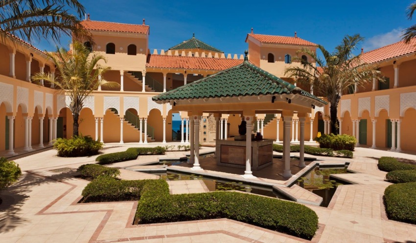 Courtyard with central fountain, surrounded by arches, palm trees, and colorful Mediterranean-style buildings under a clear blue sky.