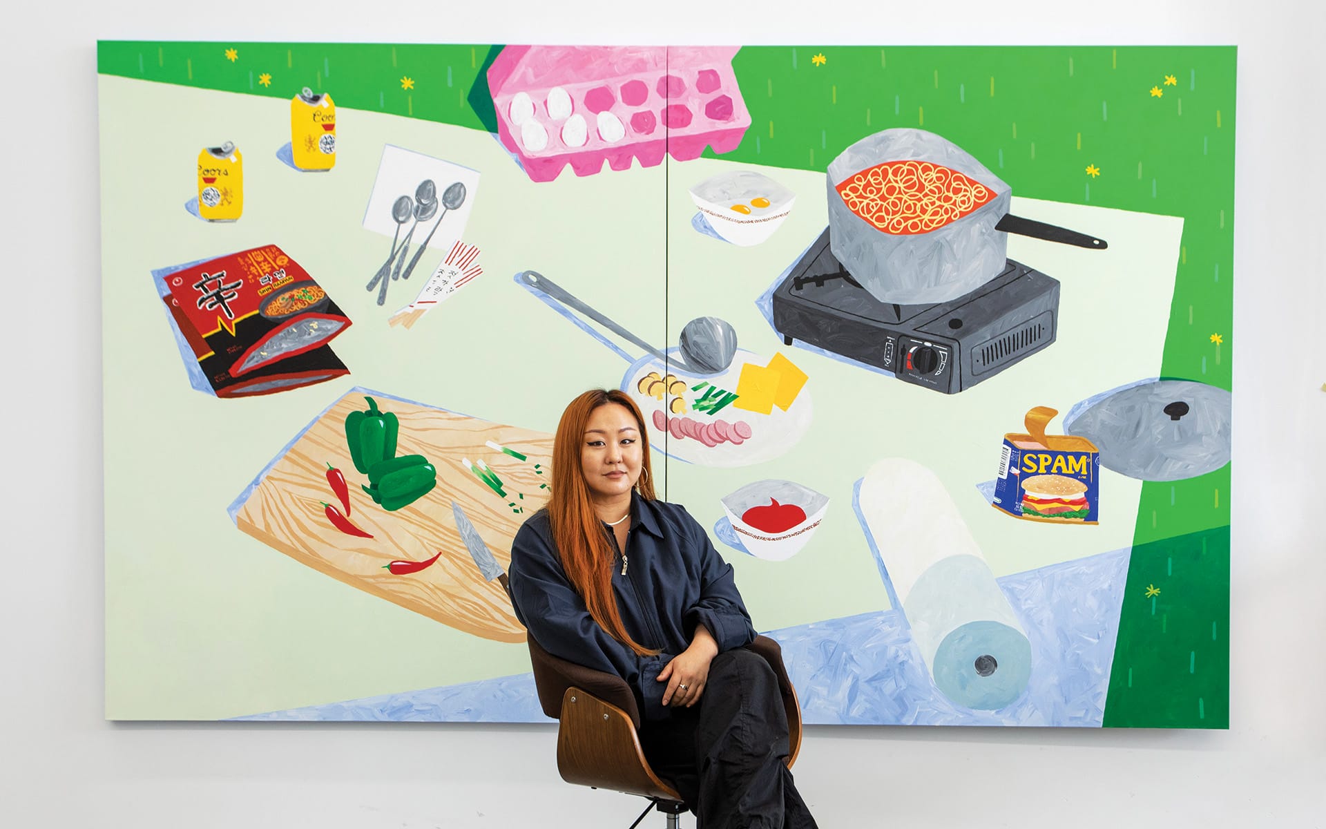 Artist sitting in front of a colorful food-themed painting with ingredients and kitchen items displayed.