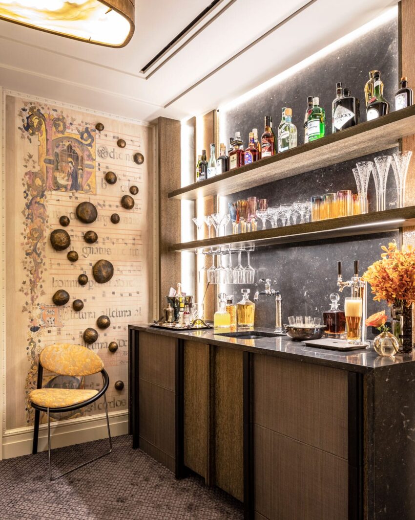 Home bar with shelves stocked with liquor bottles and glassware, a small countertop, and decorative wall art and chair.