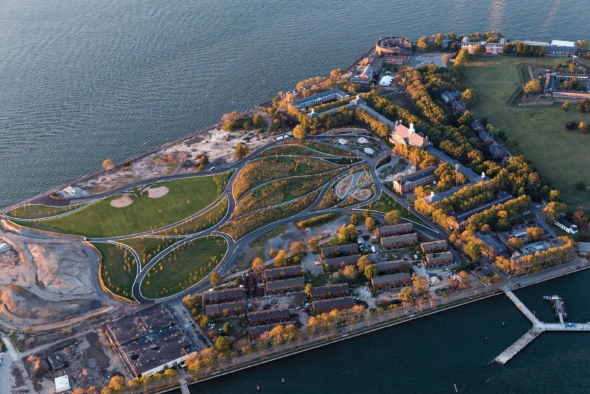 Aerial view of a green park with pathways near a waterfront, surrounded by buildings and trees.