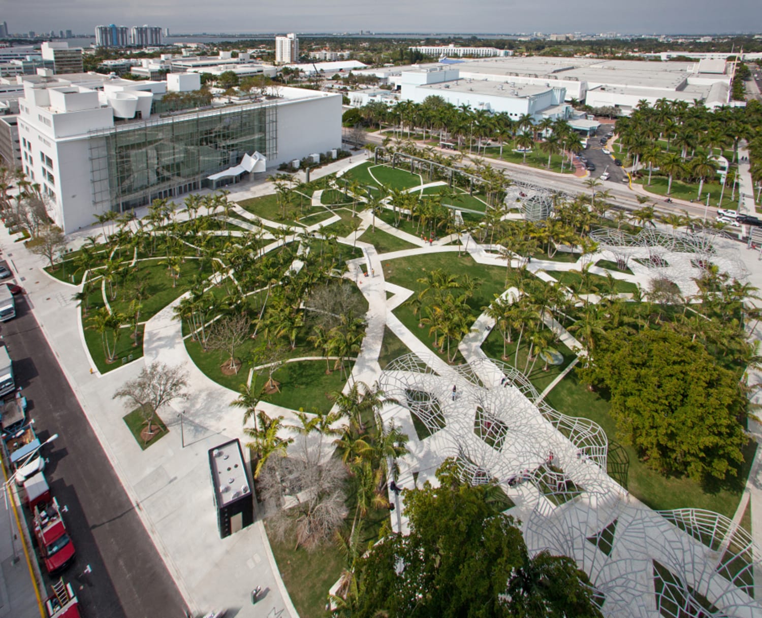 Aerial view of a modern urban park with geometric walkways, palm trees, and adjacent white building in a city setting.