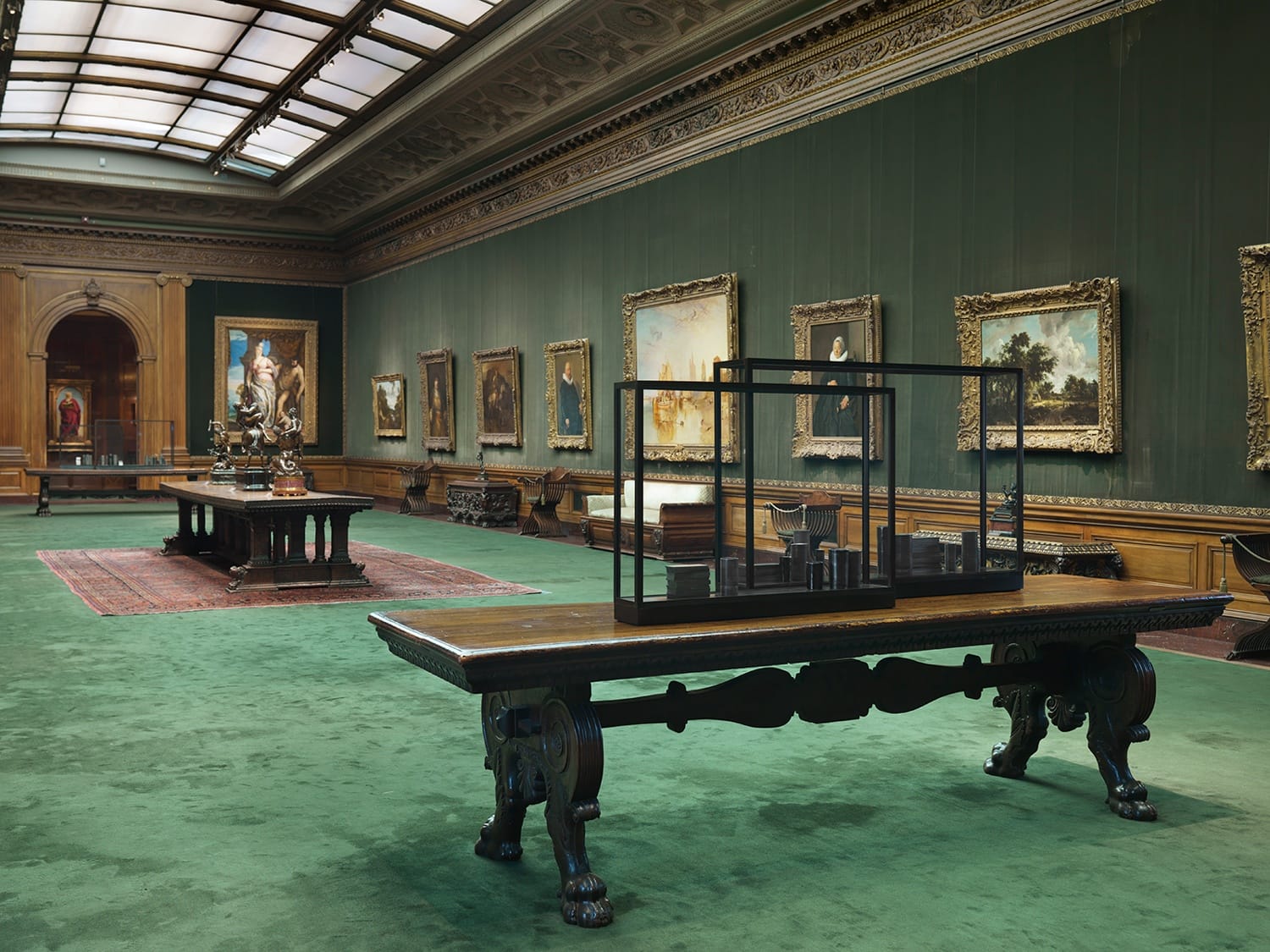 Spacious art gallery with green walls and carpet, featuring framed paintings and ornate wooden tables with displays.