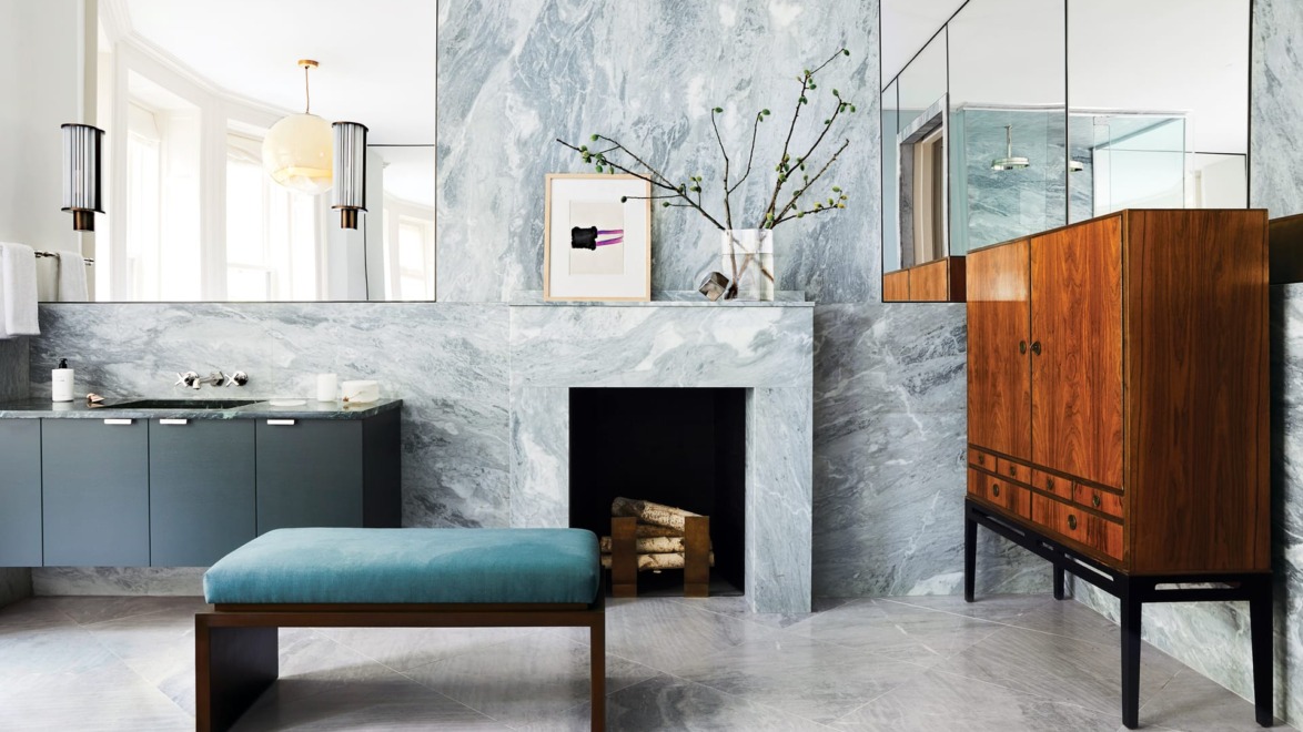 Modern bathroom with marble walls, teal bench, wooden cabinet, and a mirror above a small fireplace with minimalistic decor.