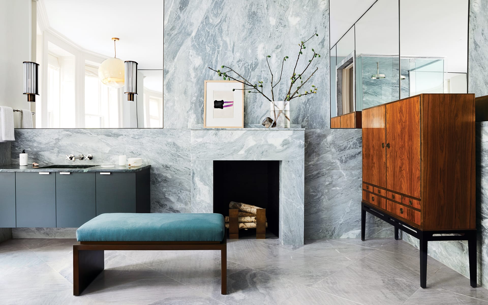 Modern bathroom with marble walls, teal bench, wooden cabinet, and a mirror above a small fireplace with minimalistic decor.