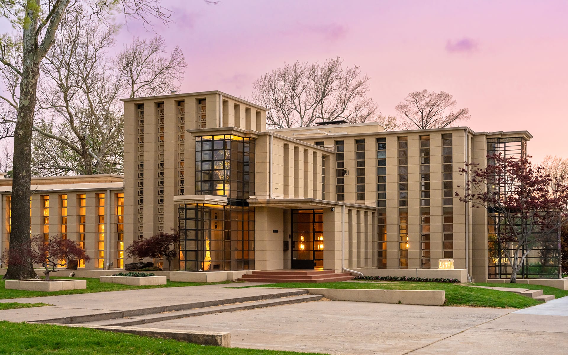 Modern architectural building with large windows, surrounded by trees and illuminated at sunset.
