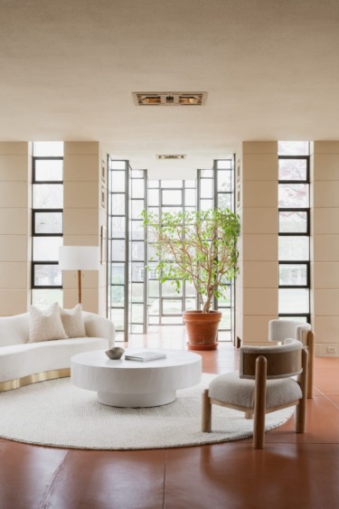 Modern living room with white furniture, a round coffee table, large potted plant, and sunlit windows.