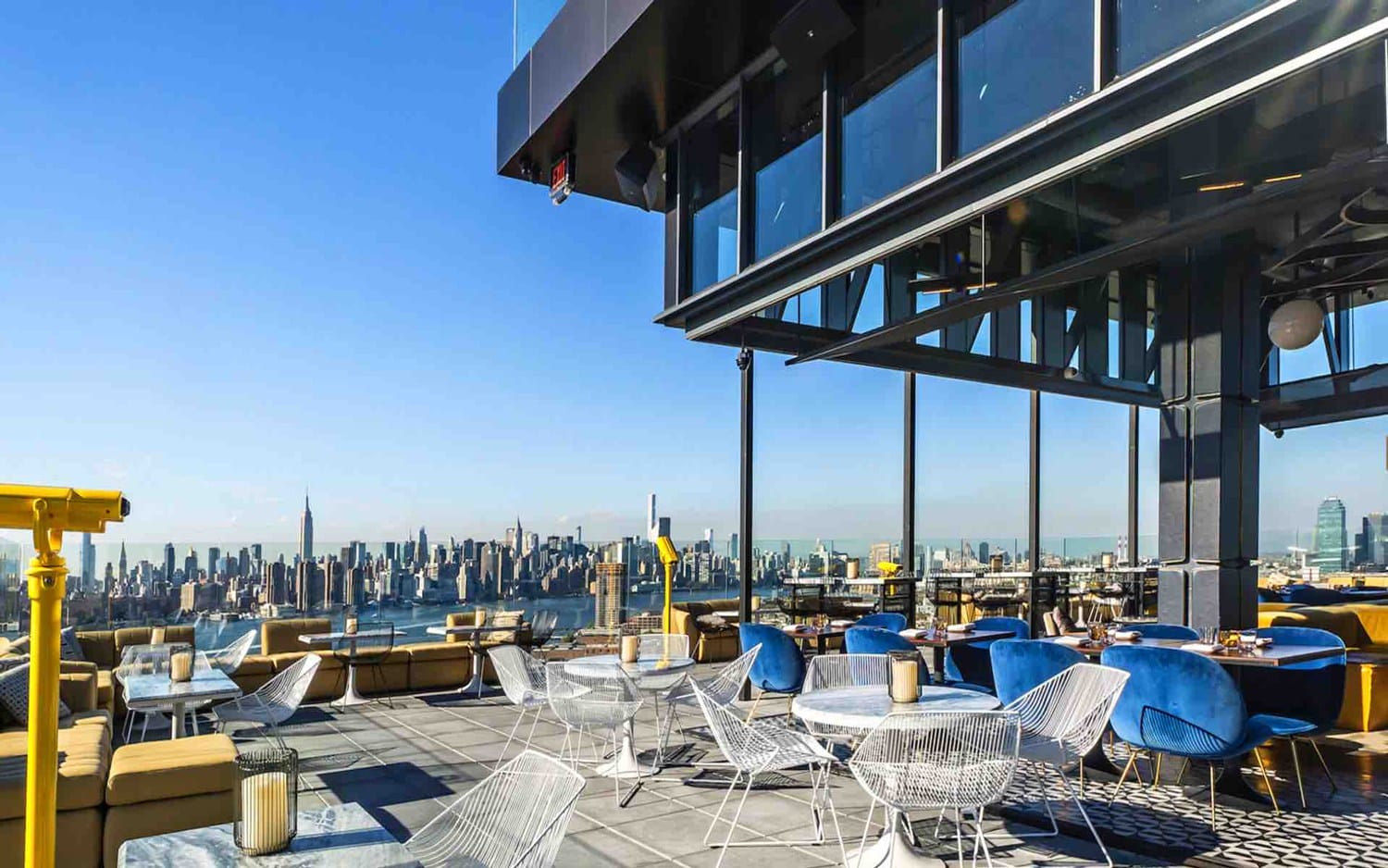 Rooftop bar with modern furniture overlooking a city skyline on a clear day.