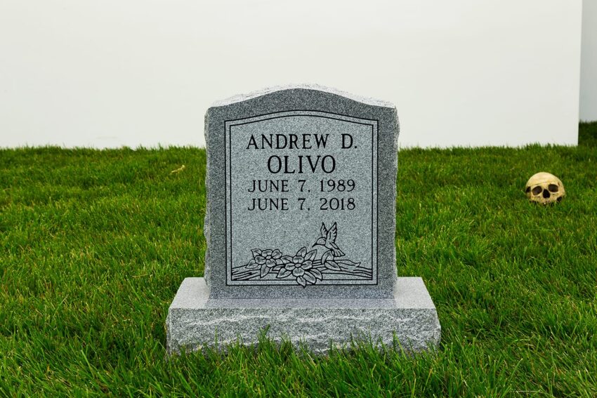 Gray tombstone on grass with engraved name Andrew D. Olivo, dates and floral decoration, skull in background.