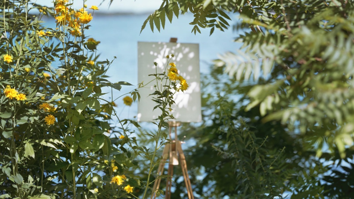 Canvas on easel surrounded by yellow flowers and green leaves near a body of water under the sunlight.