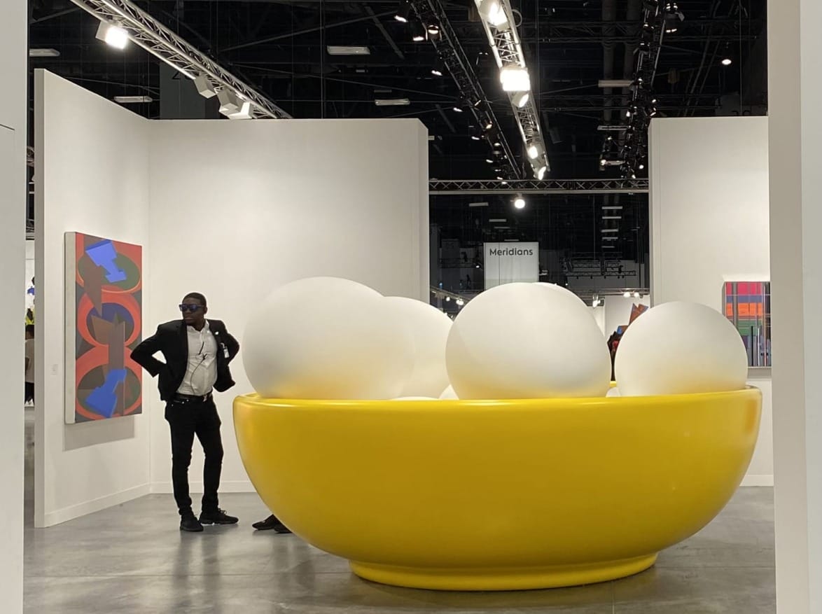Man standing by large yellow bowl filled with oversized white balls in an art gallery setting.