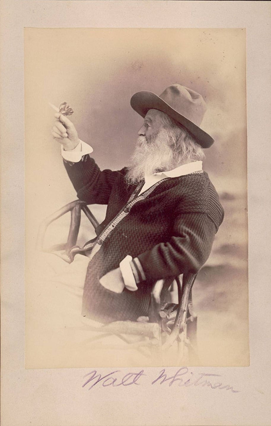 Bearded man in a hat, seated outdoors, holding a butterfly, with handwriting at the bottom of an old photograph.