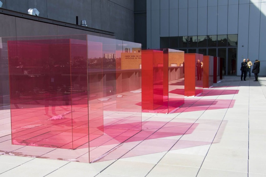 Red and pink transparent art installations on a rooftop with people observing in the background.