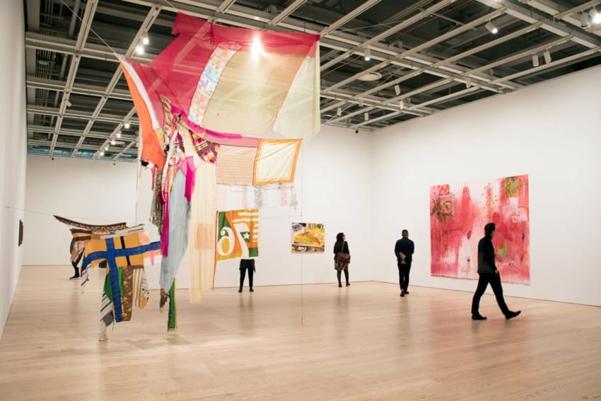 Visitors observe colorful fabric art installations in a modern gallery with wooden floors and white walls.