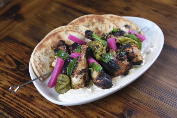 Grilled chicken kebabs on pita with pickled vegetables and leafy greens, served on a white plate on wooden table.