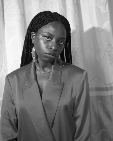 Person with braided hair wearing a suit and earrings, standing in front of a curtain. Black and white image.