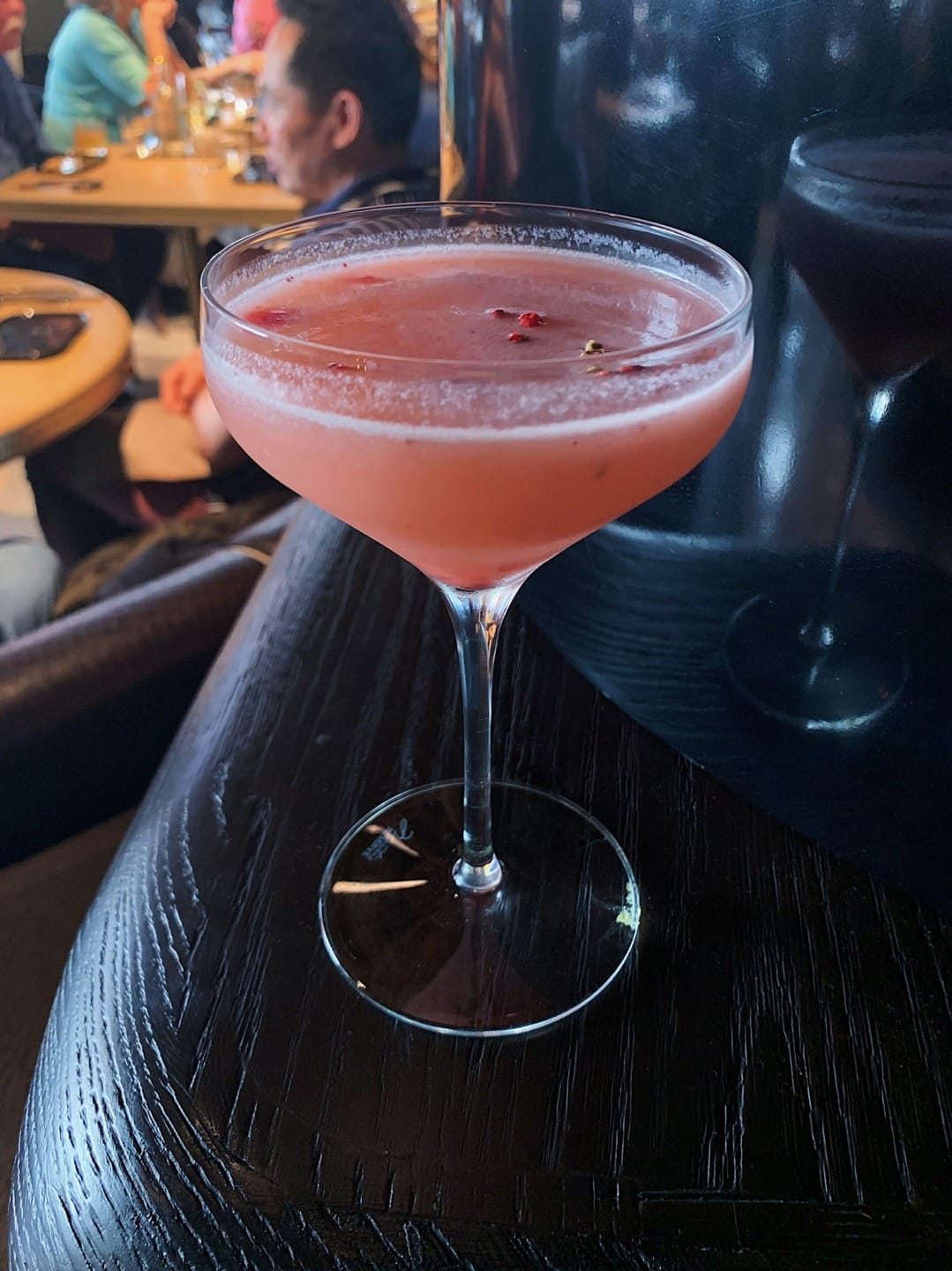 Pink cocktail in a coupe glass on a dark wooden table at a restaurant.