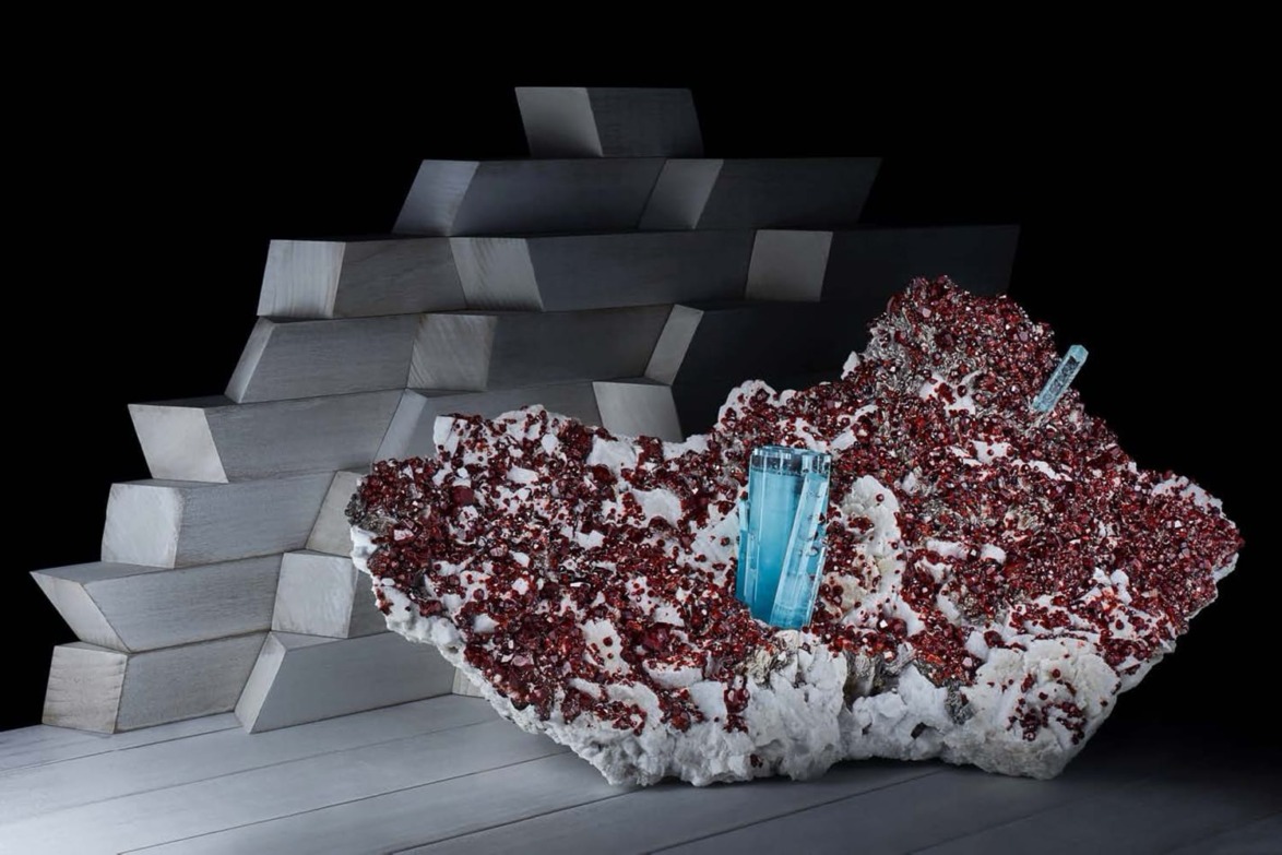A large aquamarine crystal with vibrant red garnets in a stunning mineral display with a metallic backdrop.