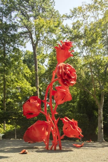 Red rose sculpture in outdoor garden setting with tall trees and greenery in the background.