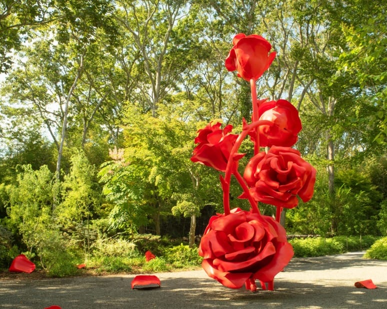 Large red rose sculpture in a garden setting with green trees and a gravel path in the background.