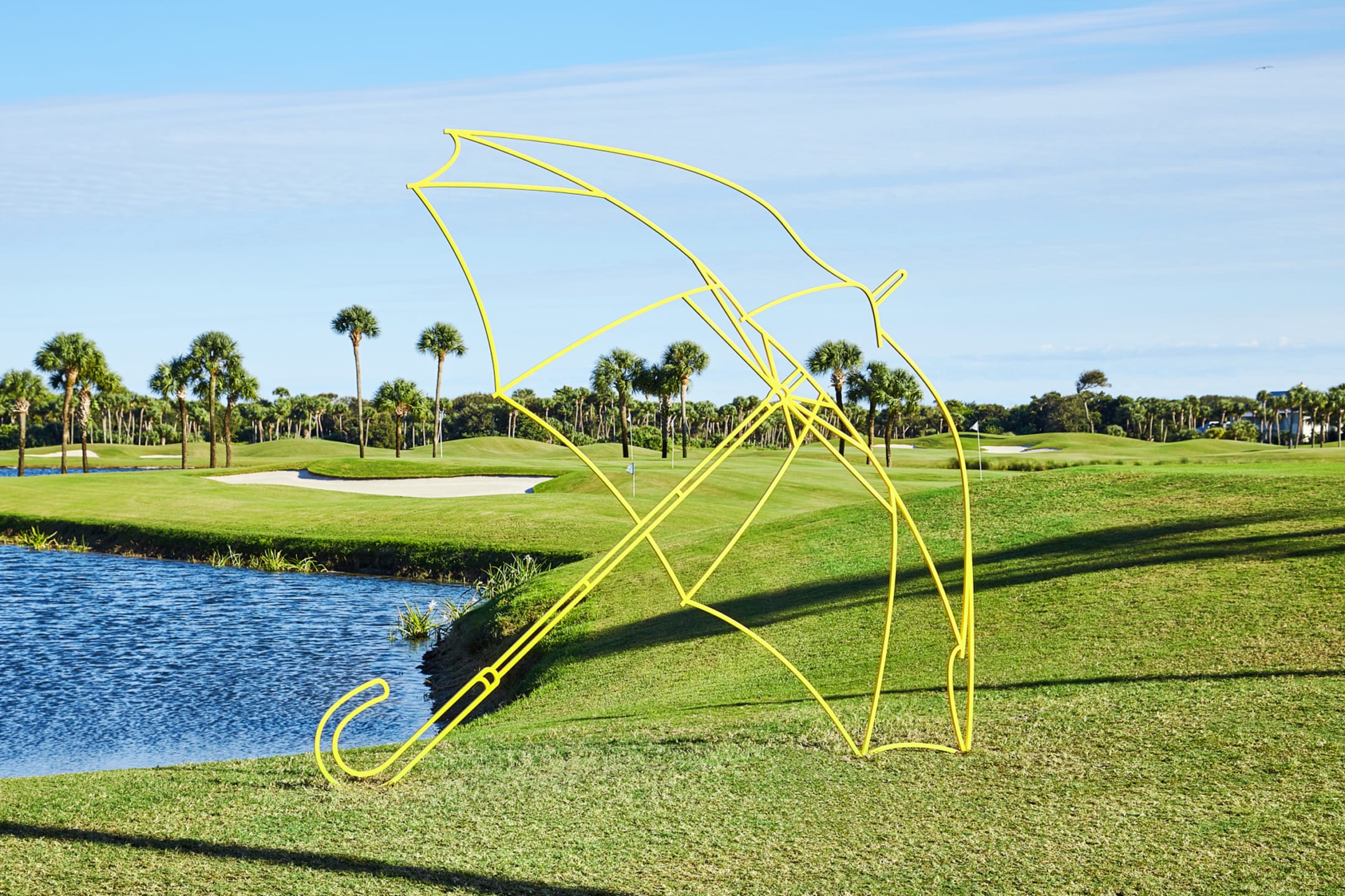 Yellow umbrella sculpture on grassy golf course with palm trees and a lake in the background under a clear blue sky.