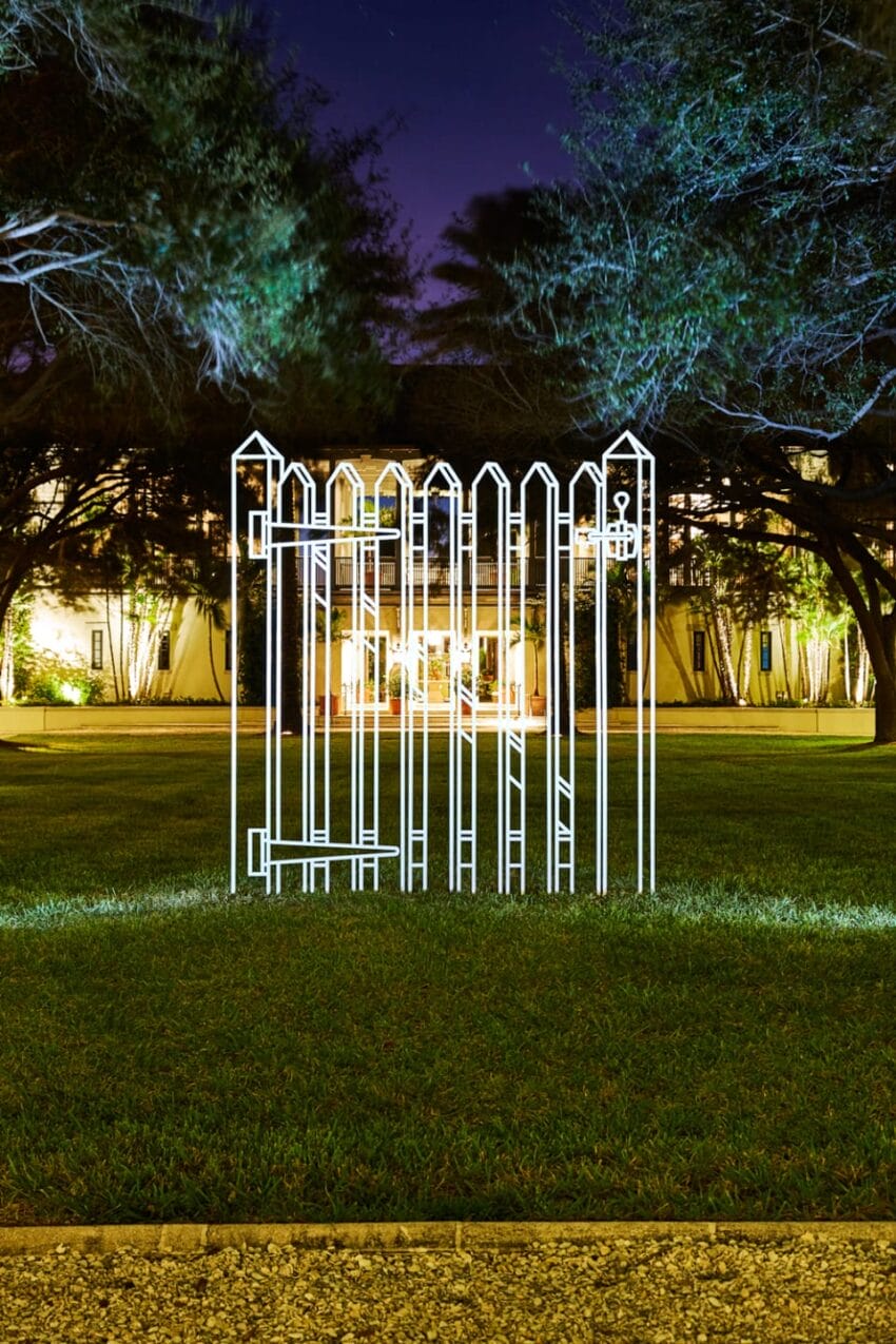 Art installation of a glowing white fence outline on a green lawn with trees and a building in the background at night.