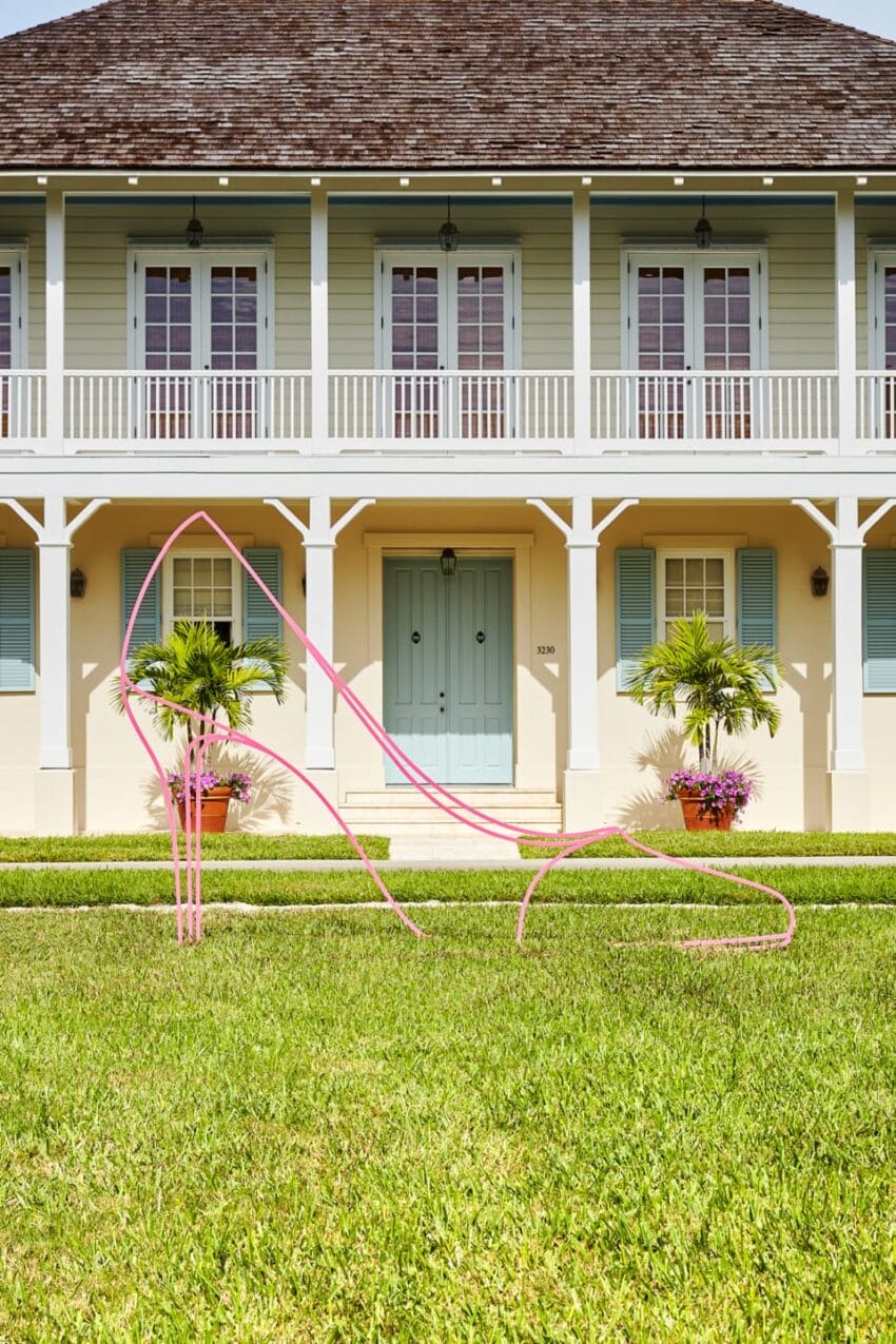 Pink high heel sculpture on green lawn in front of a two-story house with blue shutters and potted plants by the entrance.