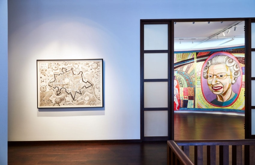 Exhibit room with framed sketch on left wall and colorful mural with older woman in background through open doorway.