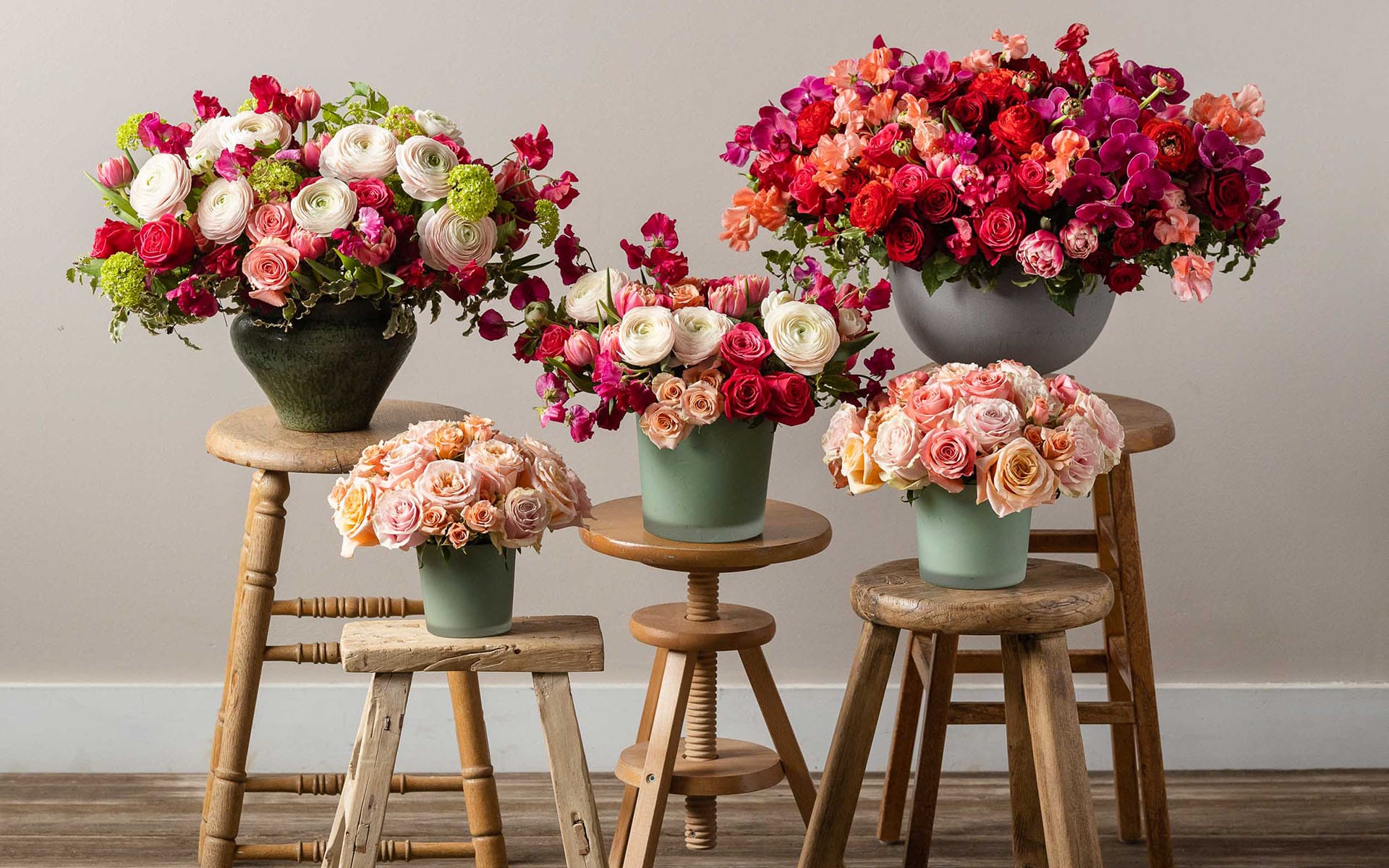 Colorful flower arrangements in various vases on wooden stools against a neutral wall background.