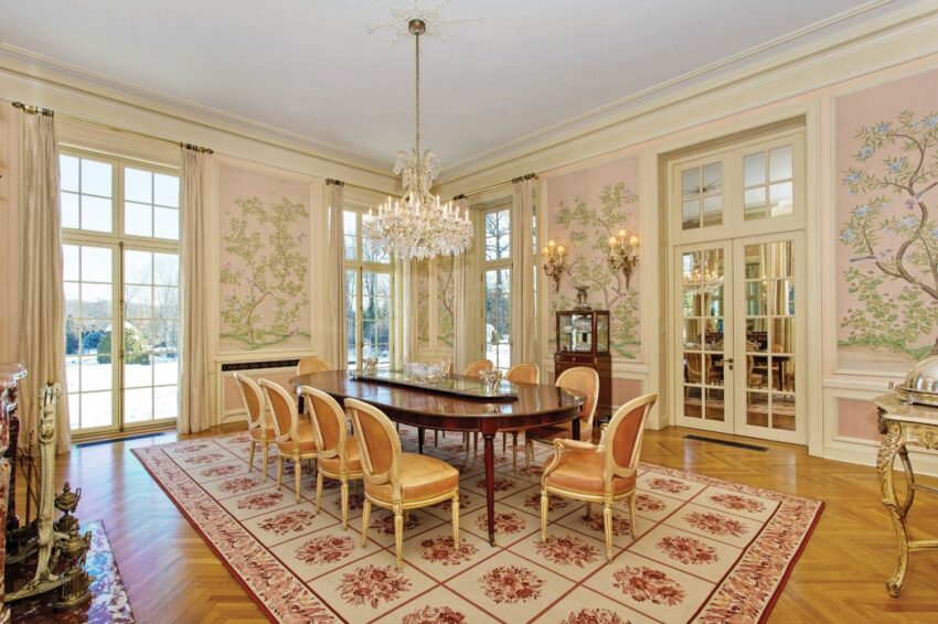 Elegant dining room with a chandelier, large windows, a long table, and light-colored chairs on a patterned rug.
