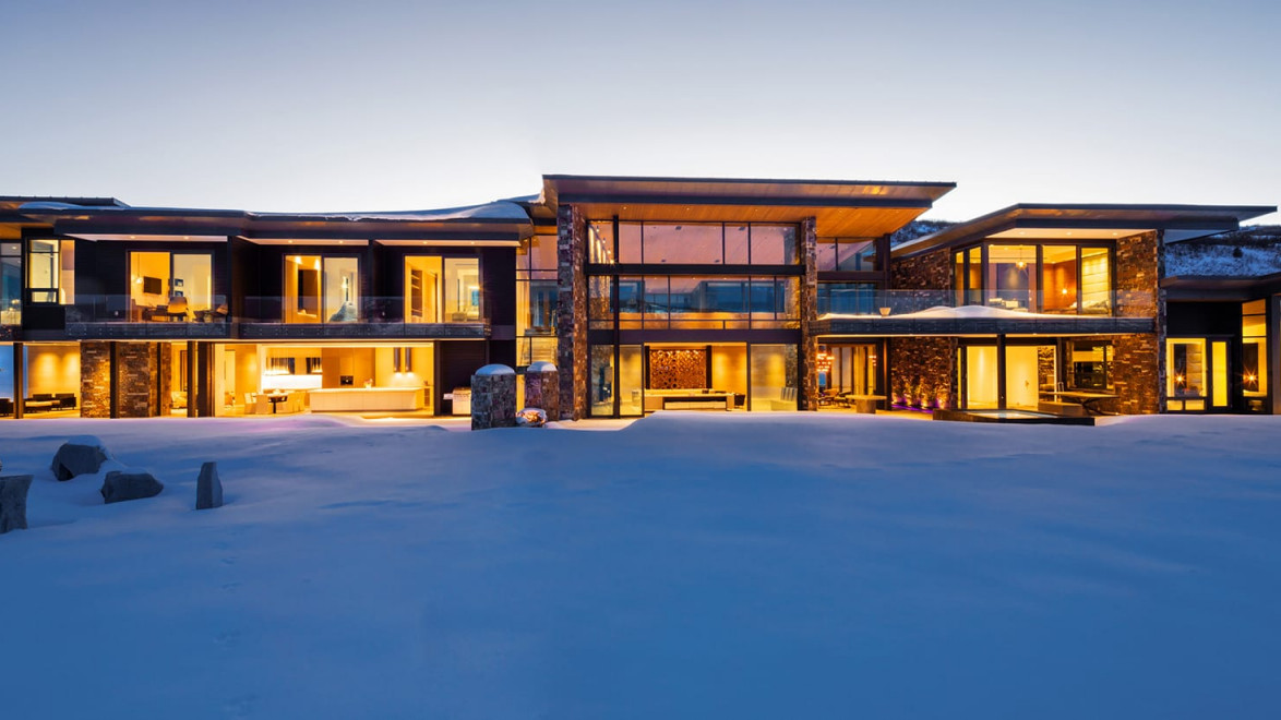 Modern glass-fronted house with warm lights, set against a snowy landscape at dusk.