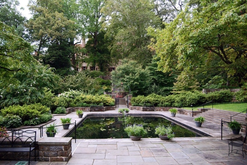 Lush garden with stone pathways, a small pond, and surrounded by trees and greenery on a serene day.
