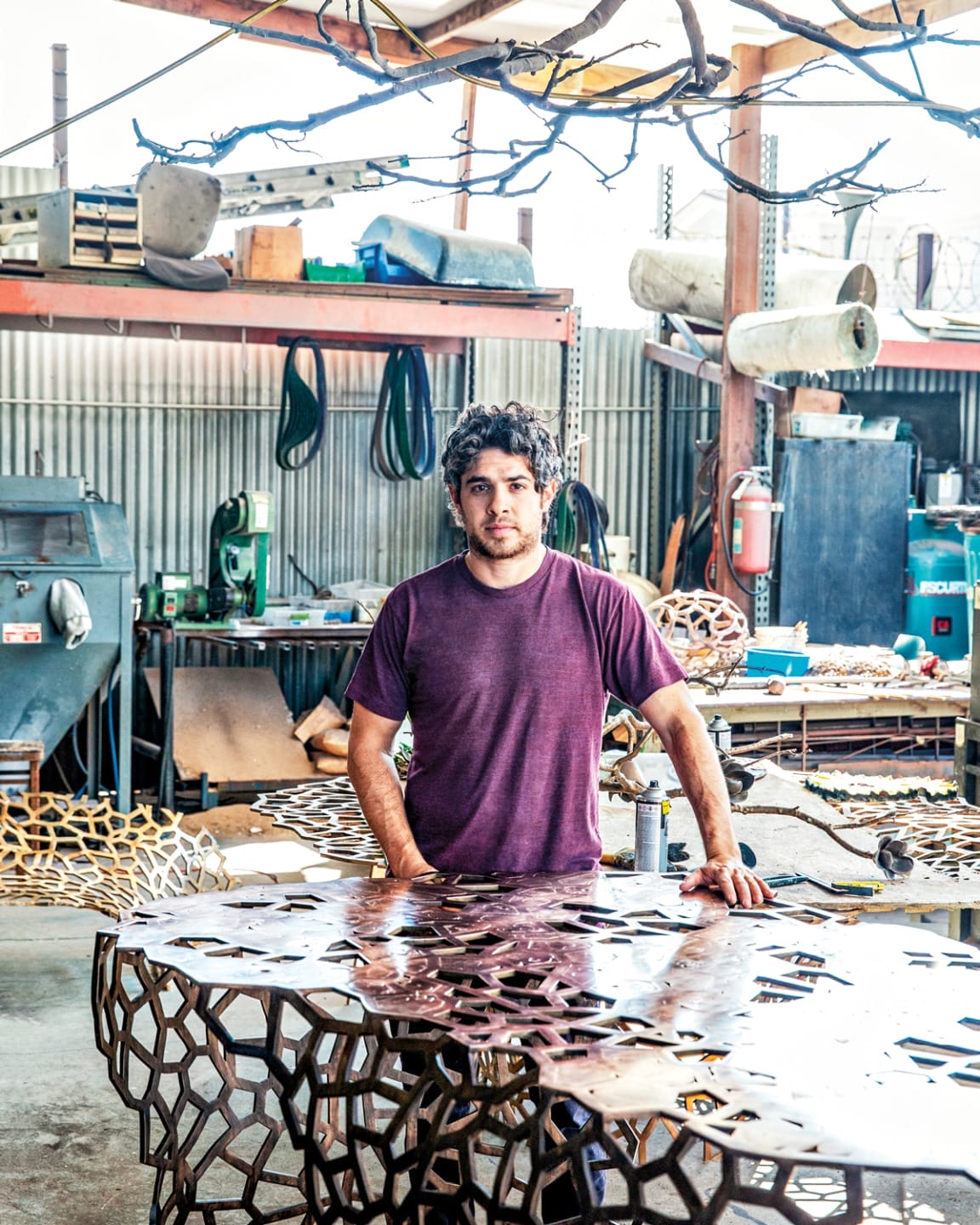 Man standing in an art studio with metal sculptures and various tools around him, wearing a purple shirt, industrial setting.