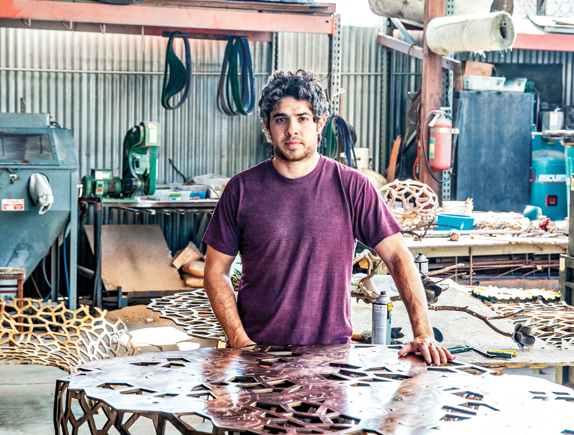 A man stands in a workshop filled with machinery, surrounded by metal artworks, wearing a maroon shirt.