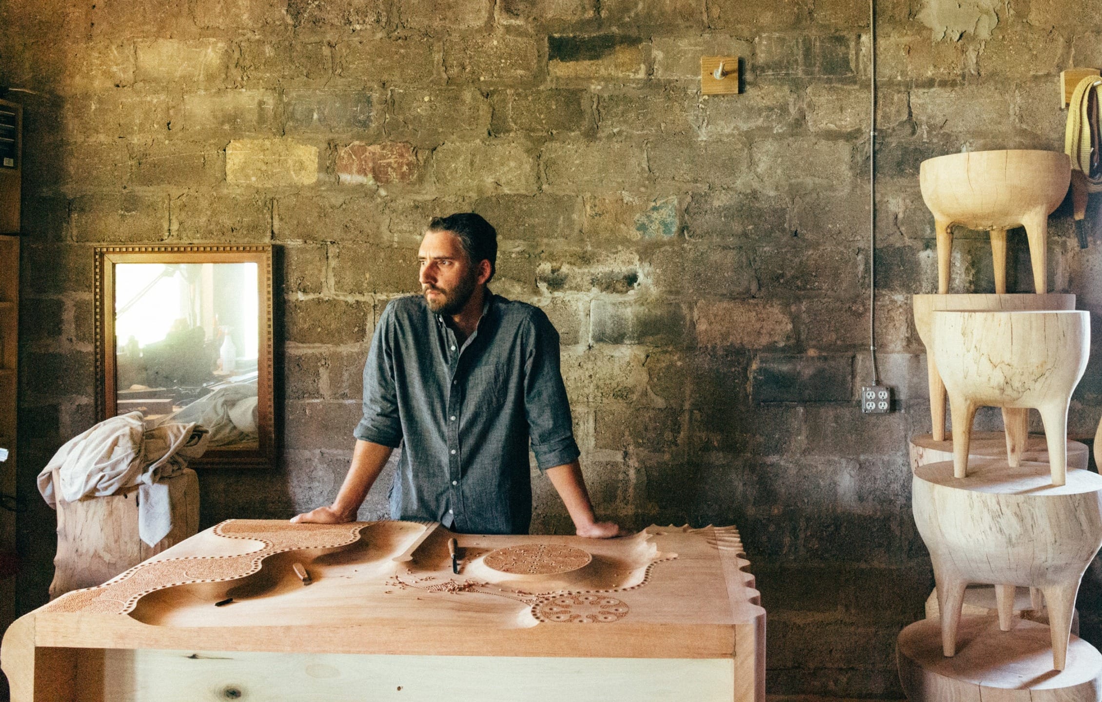 Man standing in a workshop with a carved wooden table in front and stack of wooden furniture on the side.