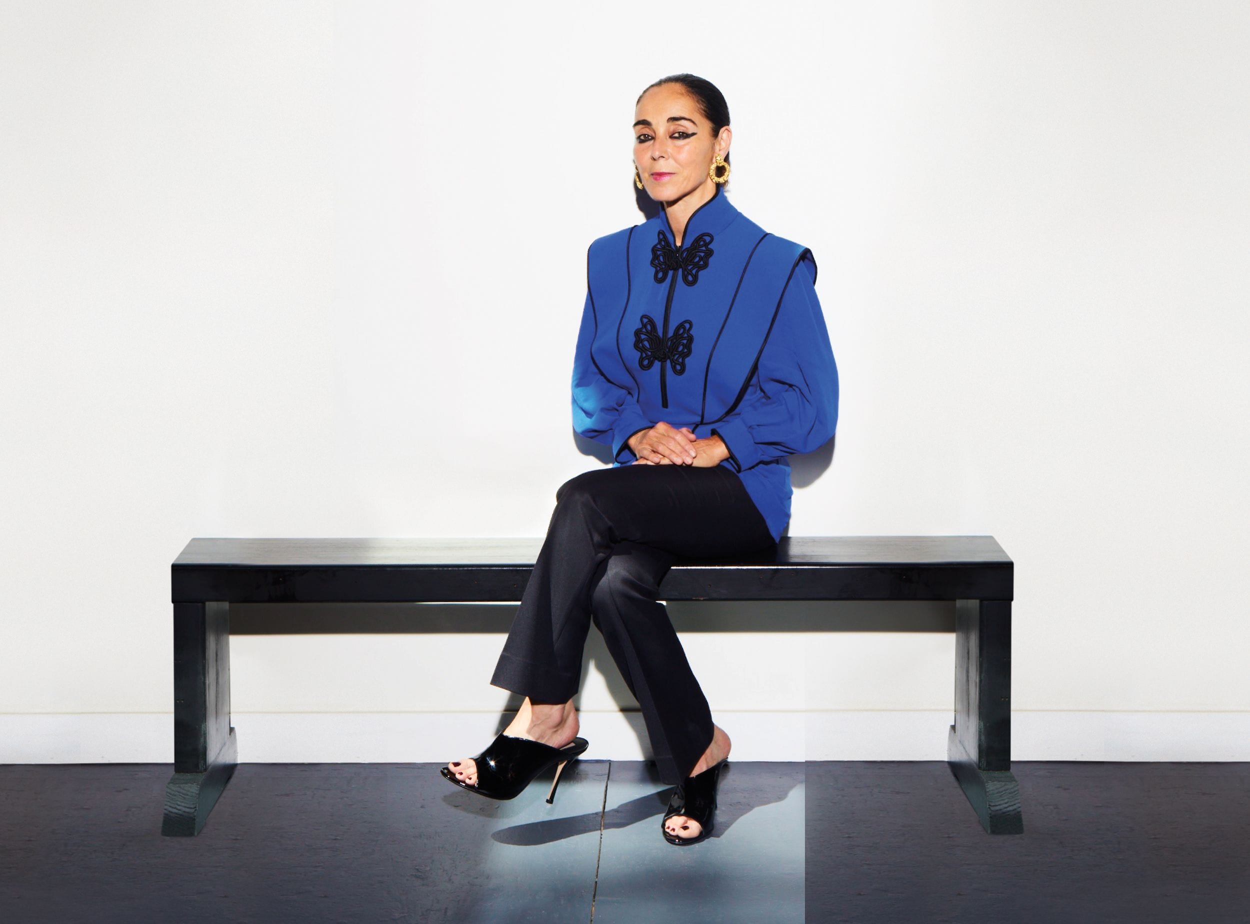 Person sitting on a bench wearing a blue top and black pants, with a white wall background.