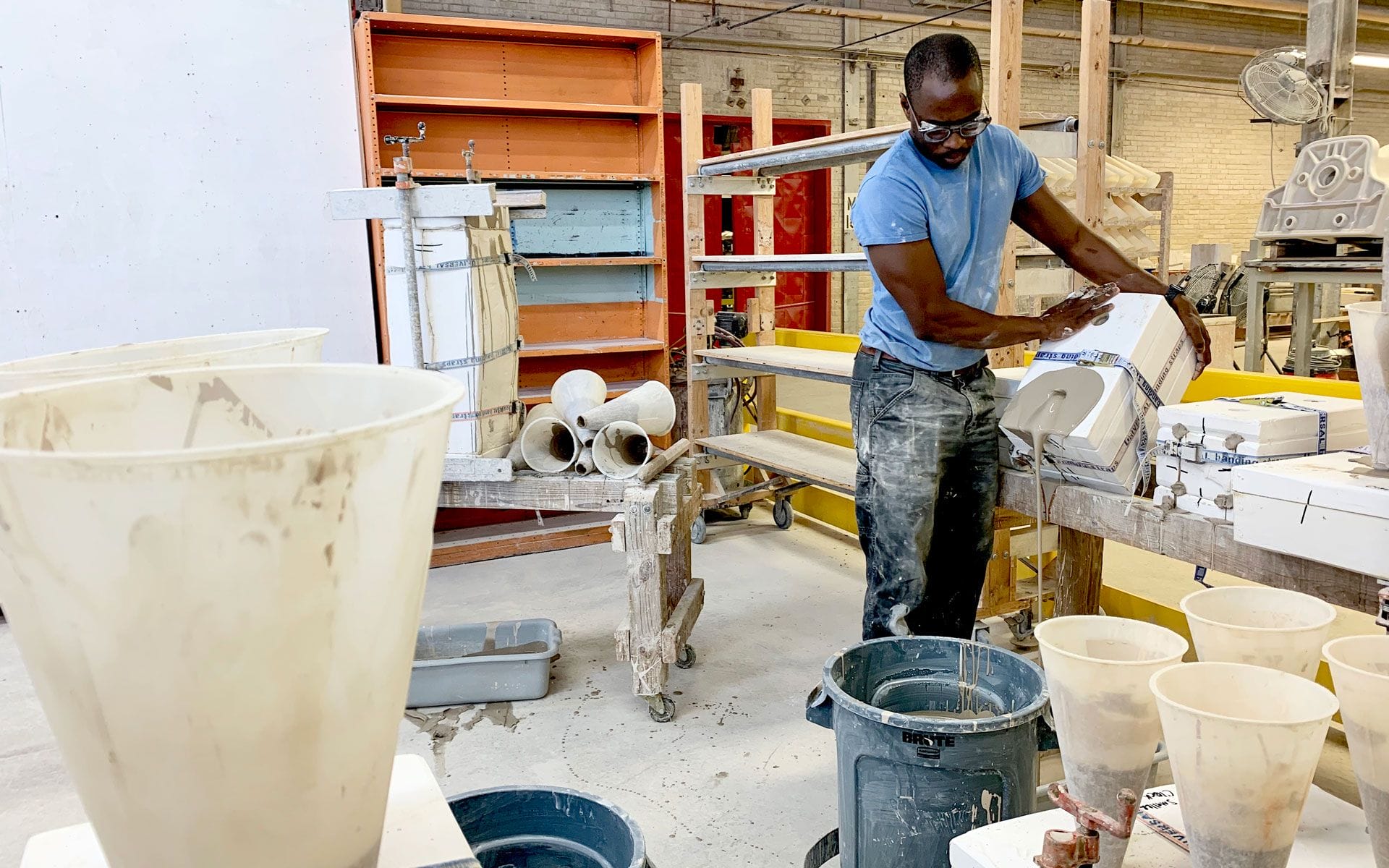 Man working in a ceramics studio, surrounded by shelves, tools, and pottery supplies.
