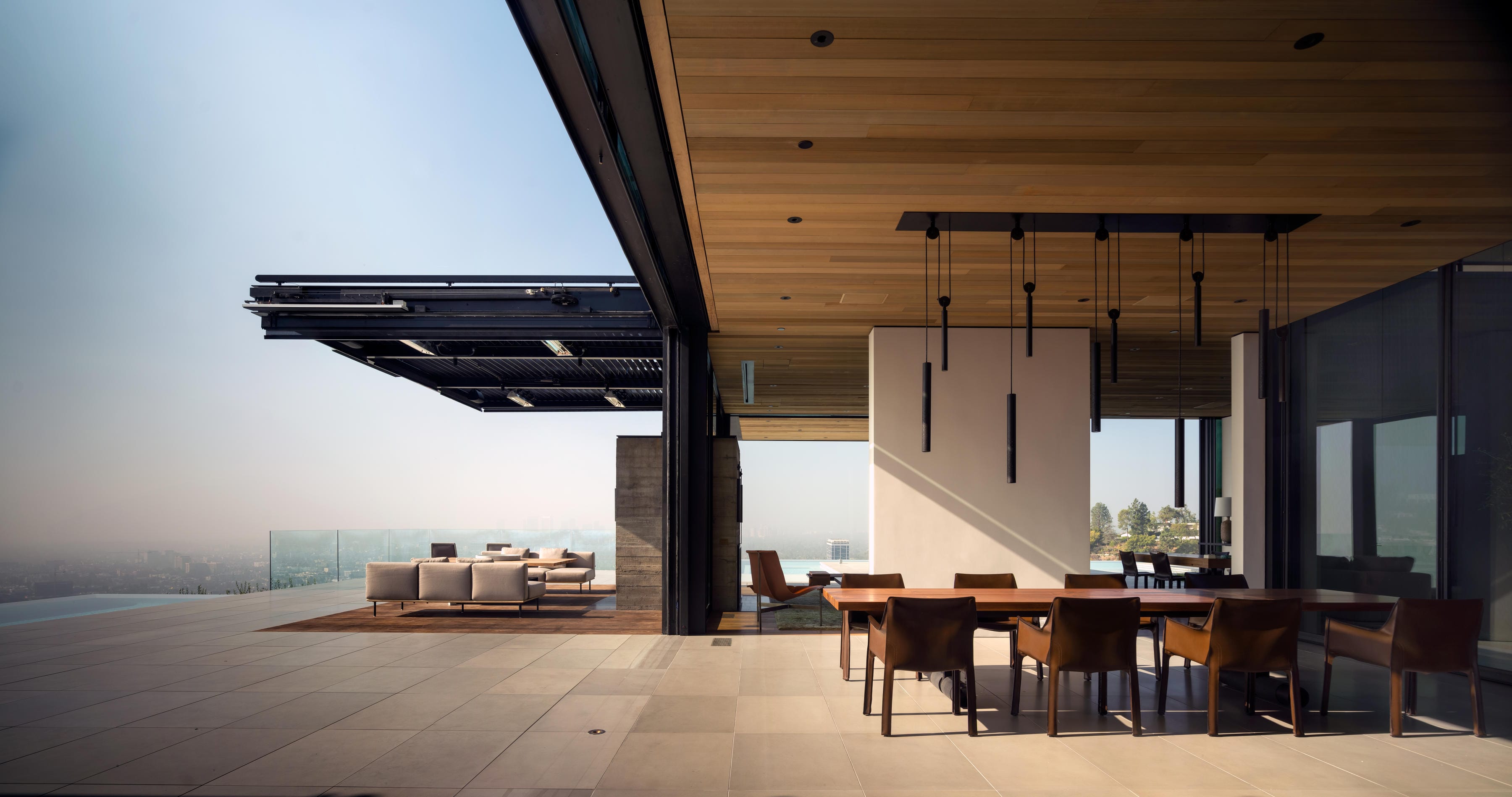 Modern open-air living space with a dining area and pool, overlooking a cityscape under a clear blue sky.