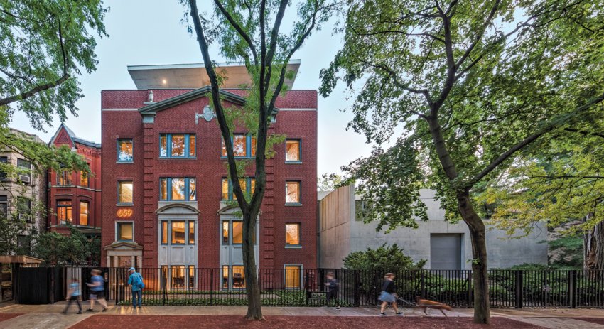 Red brick building with bay windows, surrounded by trees, evening lighting, people walking on sidewalk in front
