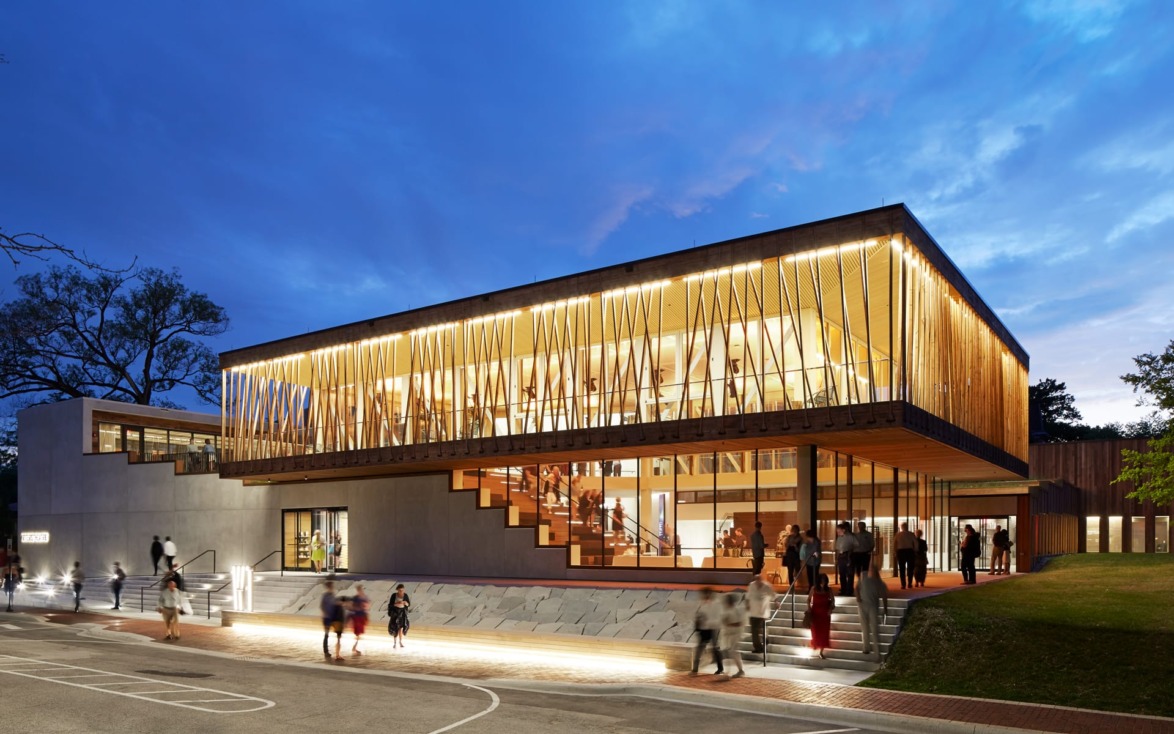 Modern illuminated building with wood accents and large windows, people walking around outside at dusk.