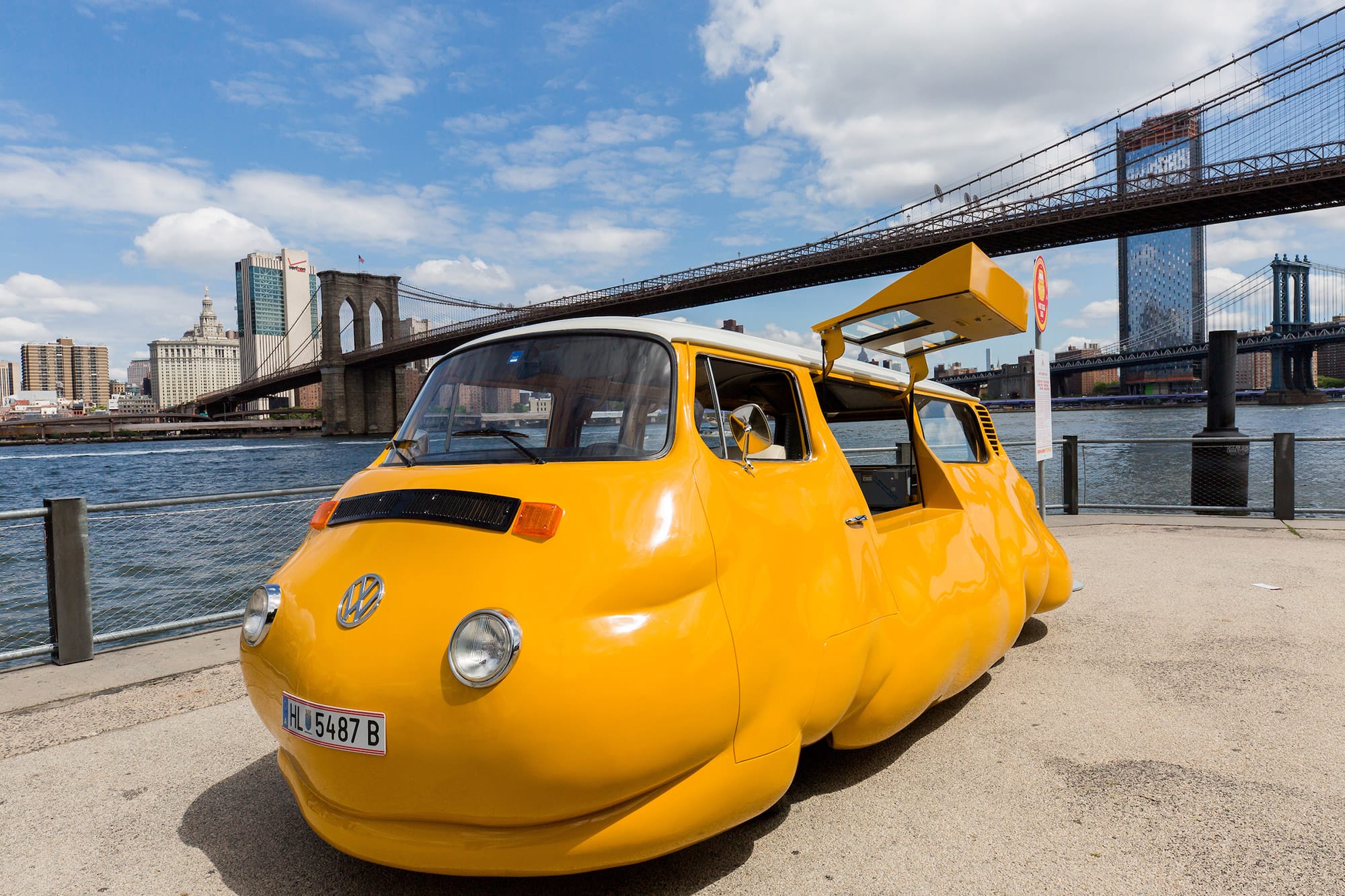 Yellow, oddly shaped vehicle parked near a river with two bridges and a cityscape in the background on a sunny day.