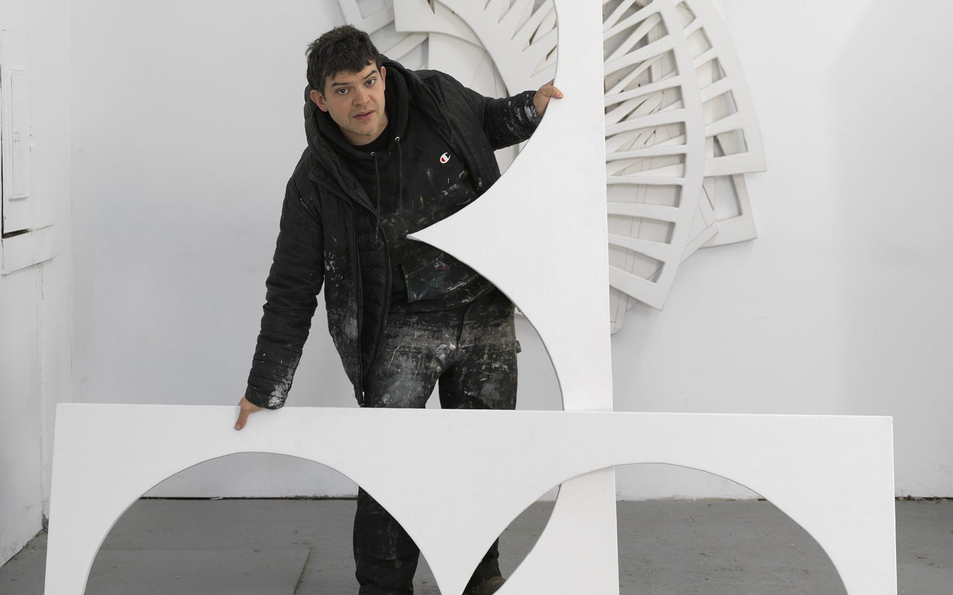 Person holding abstract geometric art pieces in a studio with a large circular sculpture displayed in the background.