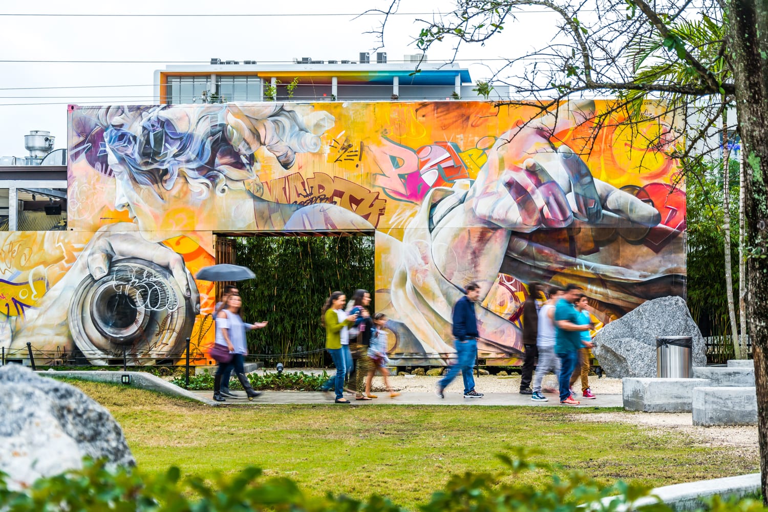Colorful graffiti mural with people walking by in a park setting, showcasing vibrant urban street art and nature.