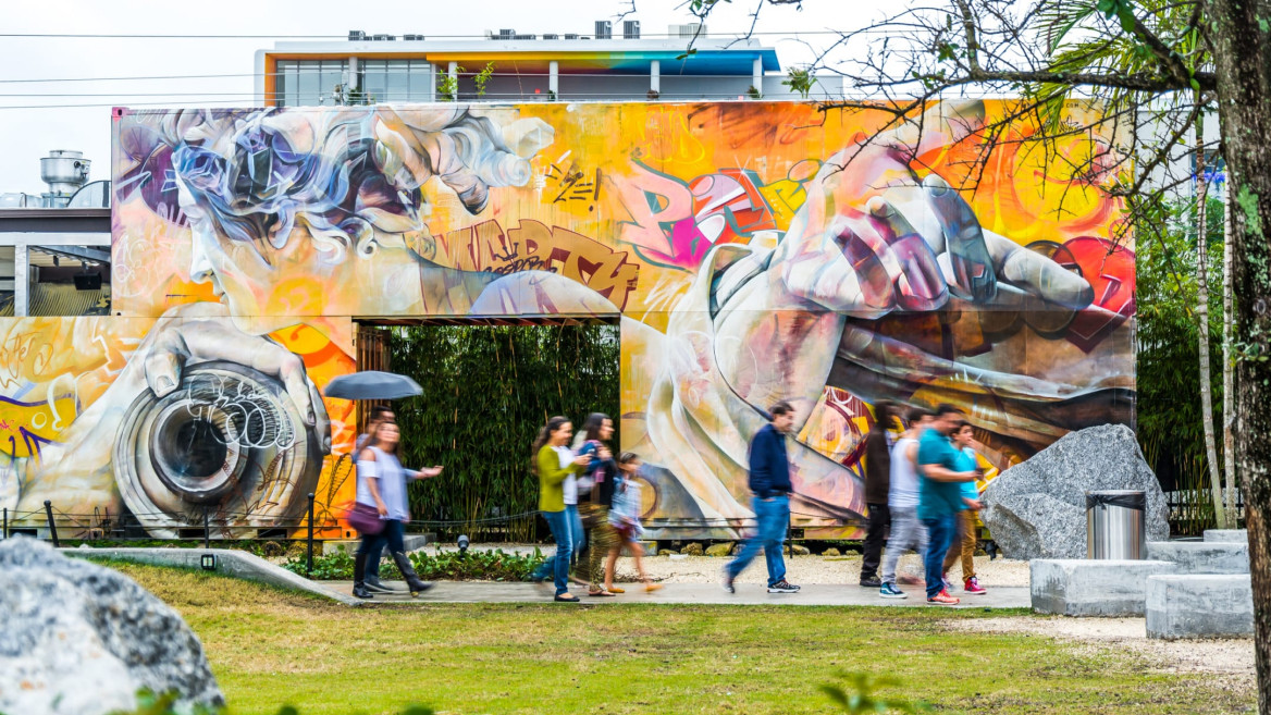 Colorful urban mural with abstract designs and people walking in front of it on a cloudy day.
